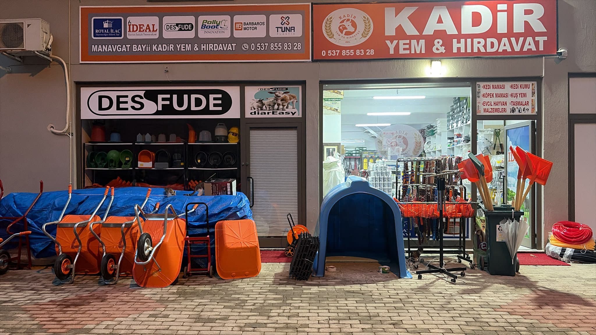 Kadir Yem & Hırdavat'ın Manavgat'taki dükkân cephesi: turuncu tabela, önde turuncu el arabaları, mavi buzağı kulübesi ve kürekler